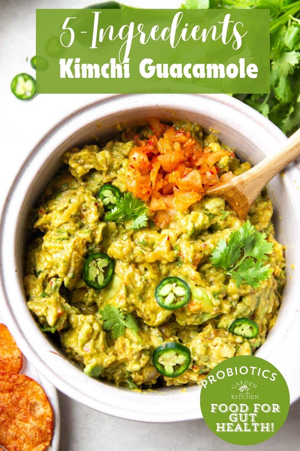 Kimchi guacamole in a white bowl with a wooden spoon, cilantro, jalapeno and chips. 