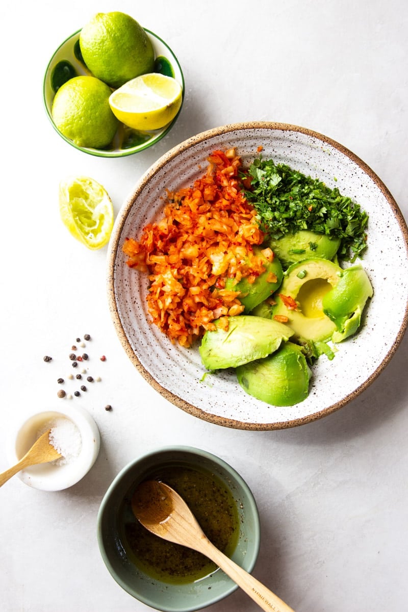 A bowl of kimchi, cilantro and avocado, bowl of limes, salt, pepper corns, liquid ingredients. 