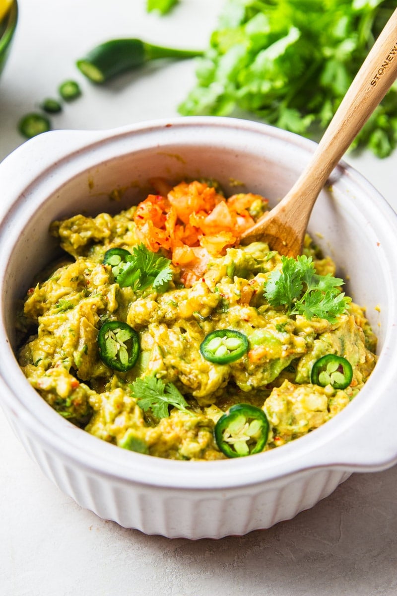 Kimchi guacamole in a white bowl with a wooden spoon, cilantro, and jalapeno.