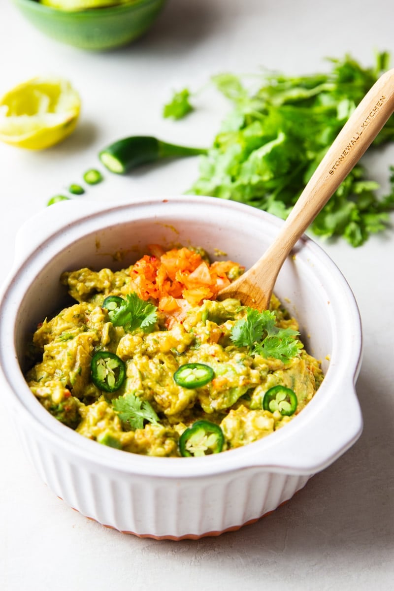 5 ingredient kimchi guacamole in a white bowl with a wooden spoon, jalapenos, cilantro, lemon.