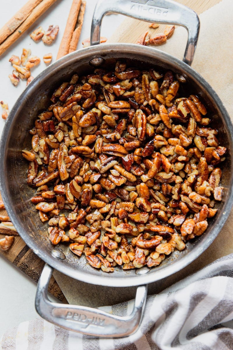 Pecans in a skillet, cinnamon sticks, pecans, and a white and black dish towel. 