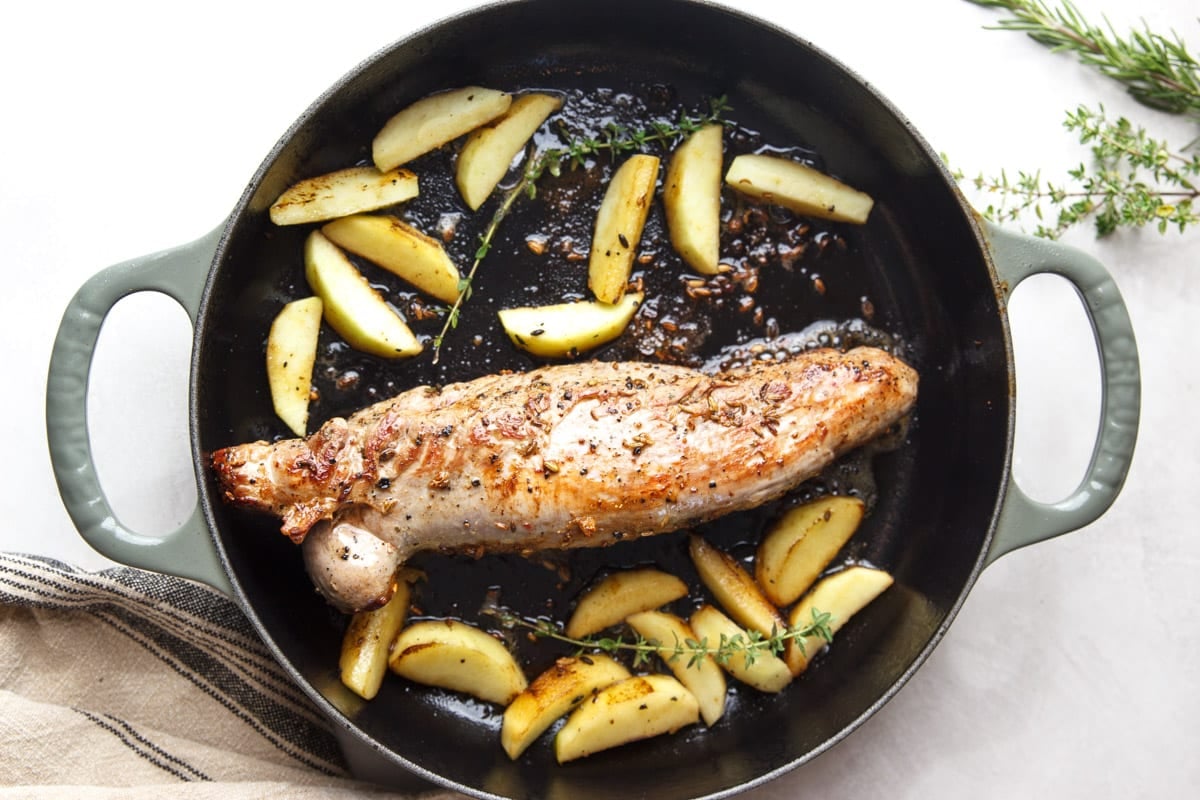 Pork tenderloin and roasted apples in a skillet with fresh thyme, and a dishtowel. 