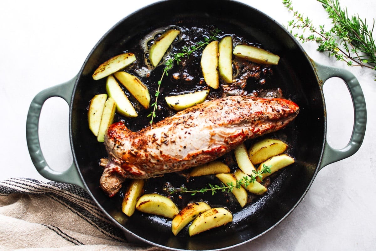 Pork tenderloin and roasted apples in a skillet with fresh thyme, and a dishtowel. 