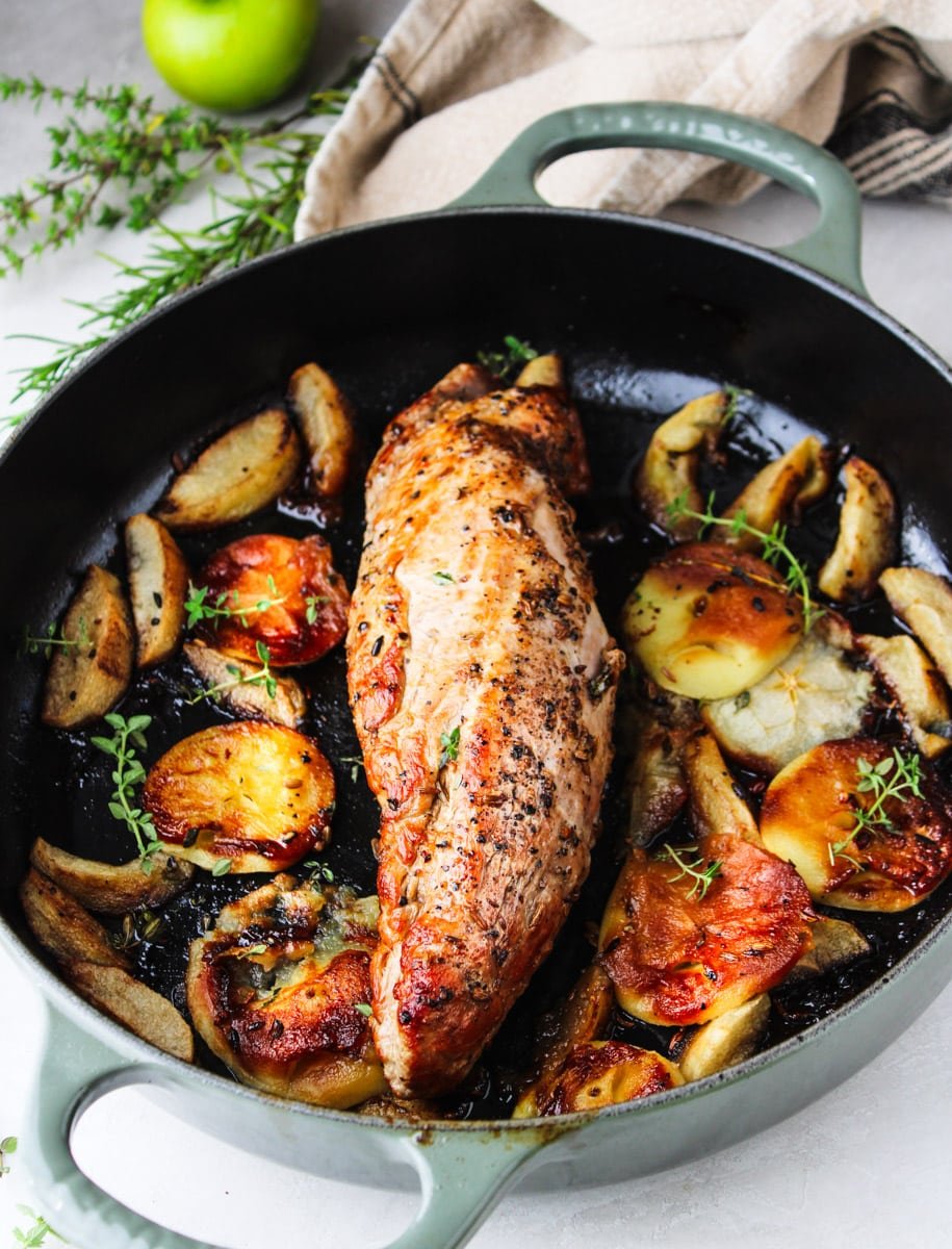 Pork tenderloin and roasted apples in a skillet with fresh thyme, a dishtowel, and a green apple. 