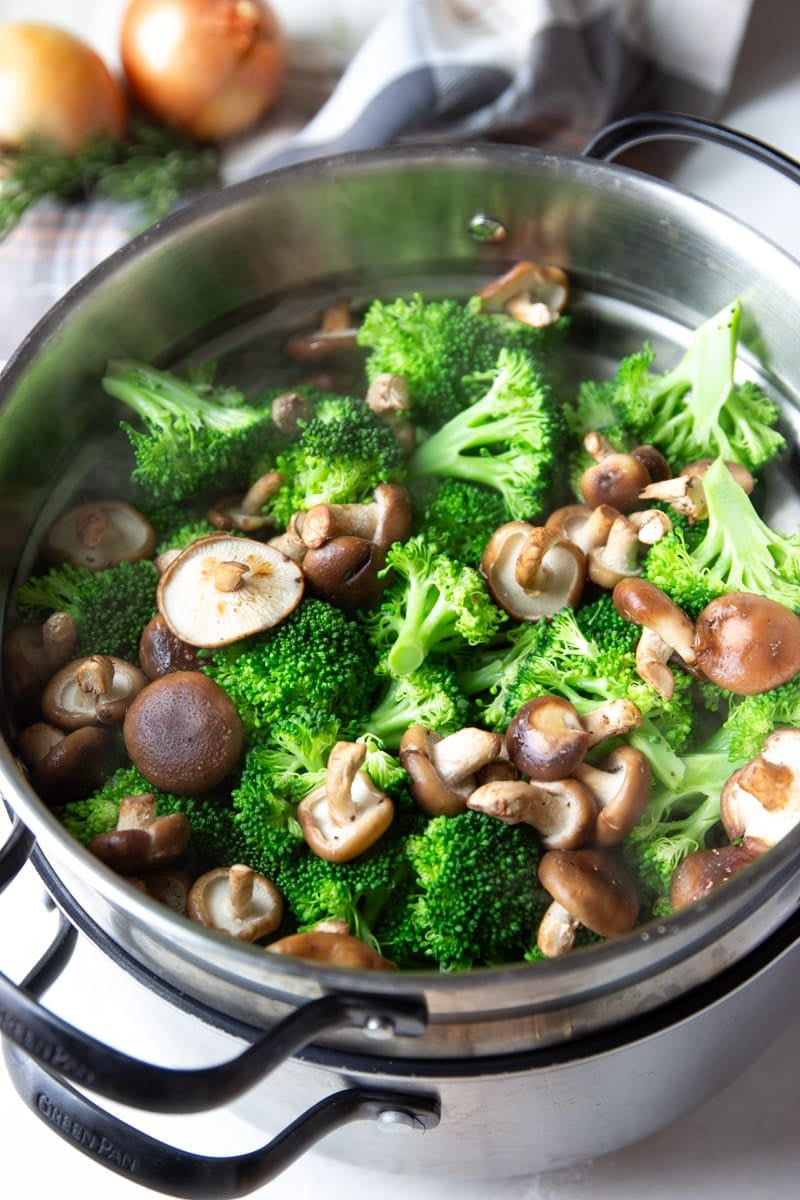 Broccoli and mushrooms in a steamer basket, onions, rosemary and plaid dish towel. 