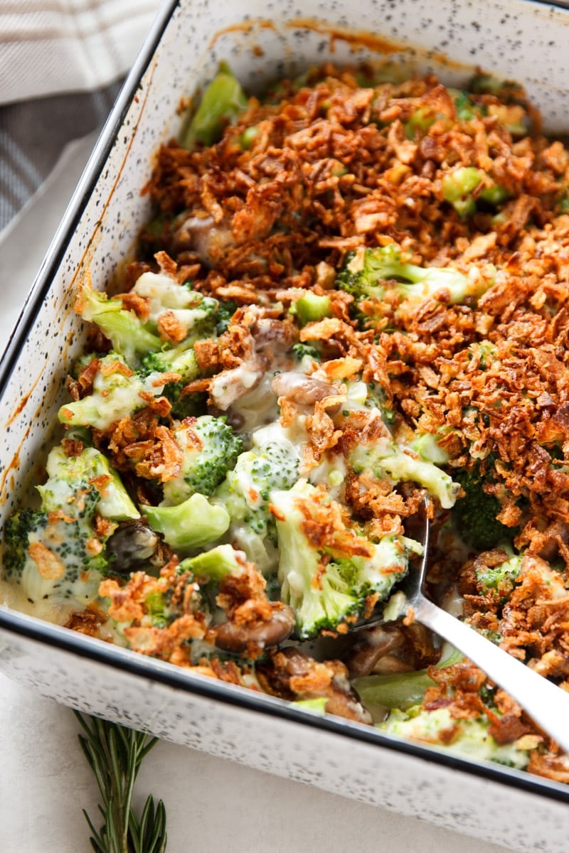 Broccoli and mushroom casserole with crispy fried onions, a silver spoon, and a plaid dish towel.