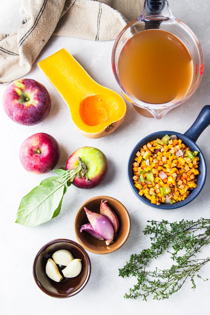 Vegetable broth in a glass measuring cup, half a butternut squash, apples,  dishtowel, chopped carrots and celery, shallots, garlic, and fresh thyme.