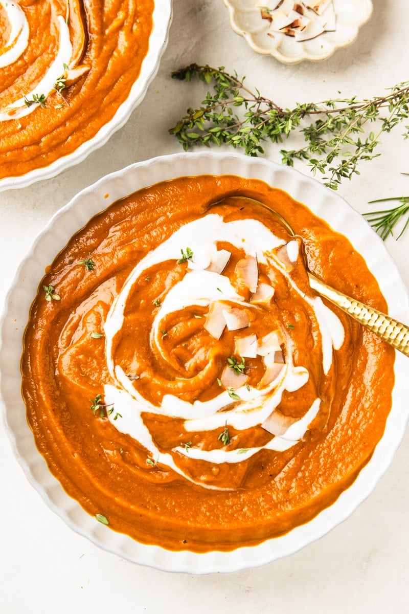 Butternut squash apple soup drizzled with yogurt and topped with coconut chips in white bowls, a white bowl of coconut chips, and fresh thyme.