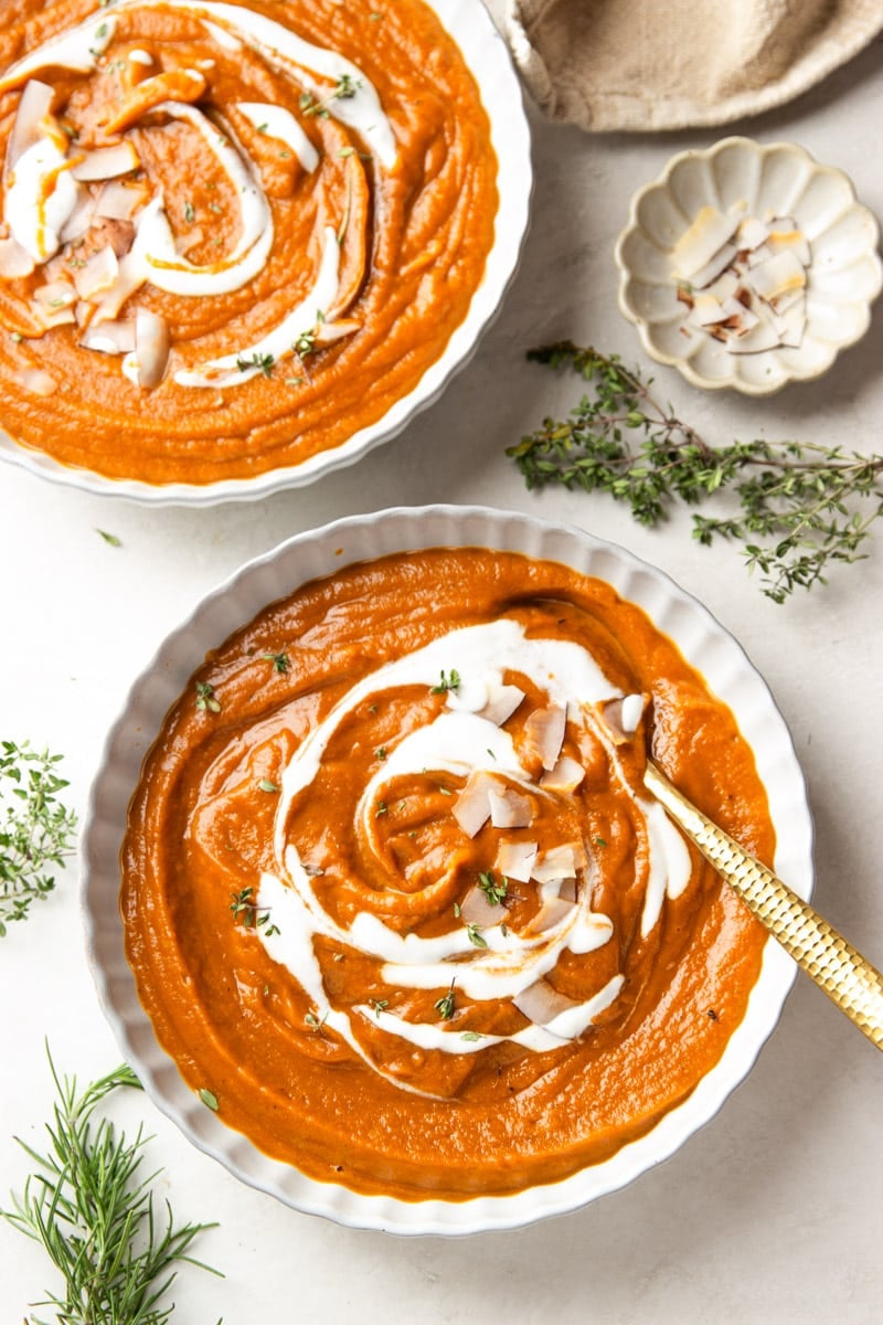 Butternut squash apple soup drizzled with yogurt and topped with coconut chips in white bowls, white bowl of coconut chips and fresh thyme.