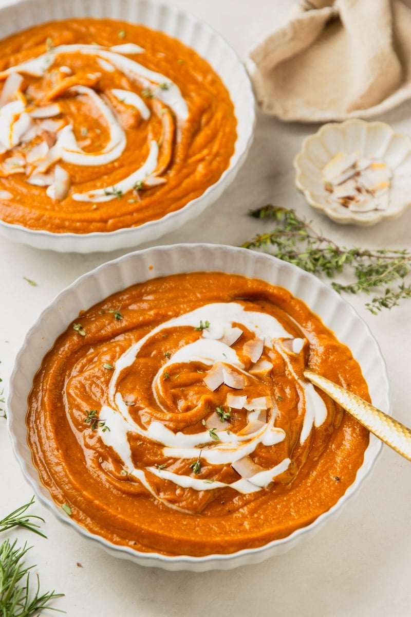 Butternut squash apple soup drizzled with yogurt and topped with coconut chips in white bowls, a white bowl of coconut chips, and fresh thyme.