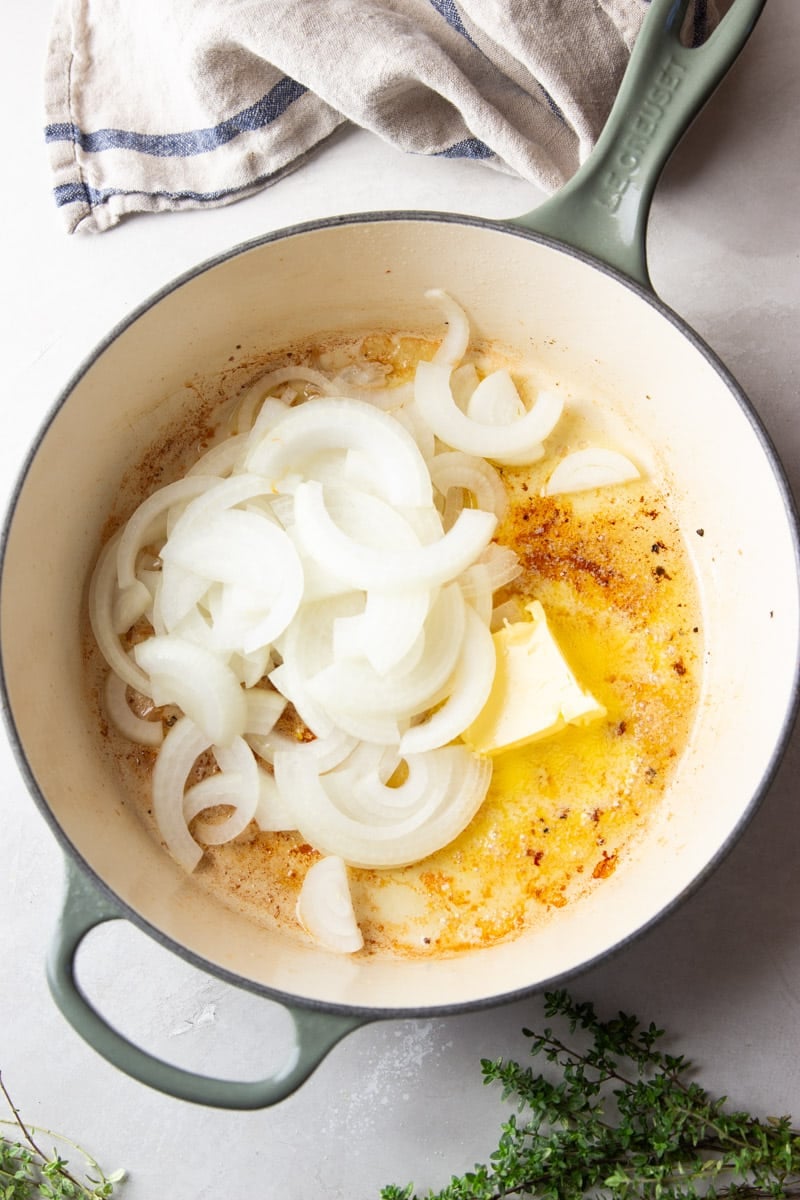 Sliced onions in a pot with butter, fresh thyme, and a striped dish towel.