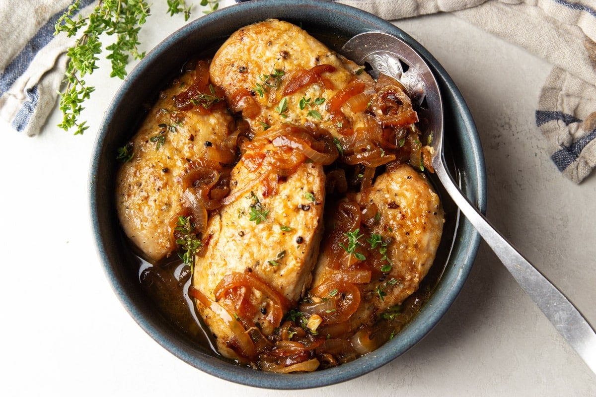 Cider braised chicken in a pot with a silver spoon, striped dish towel, and fresh thyme. 