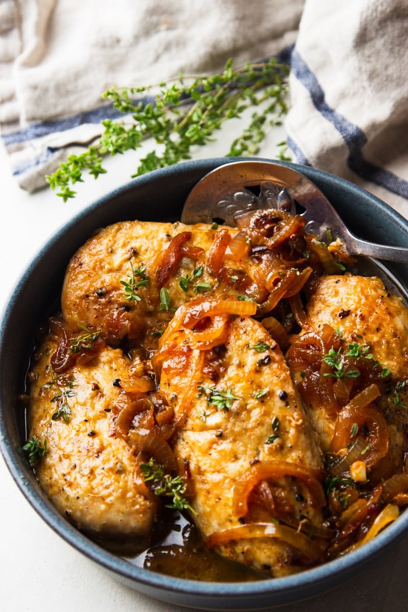 Cider braised chicken in a pot with a silver spoon, striped dish towel, and fresh thyme. 