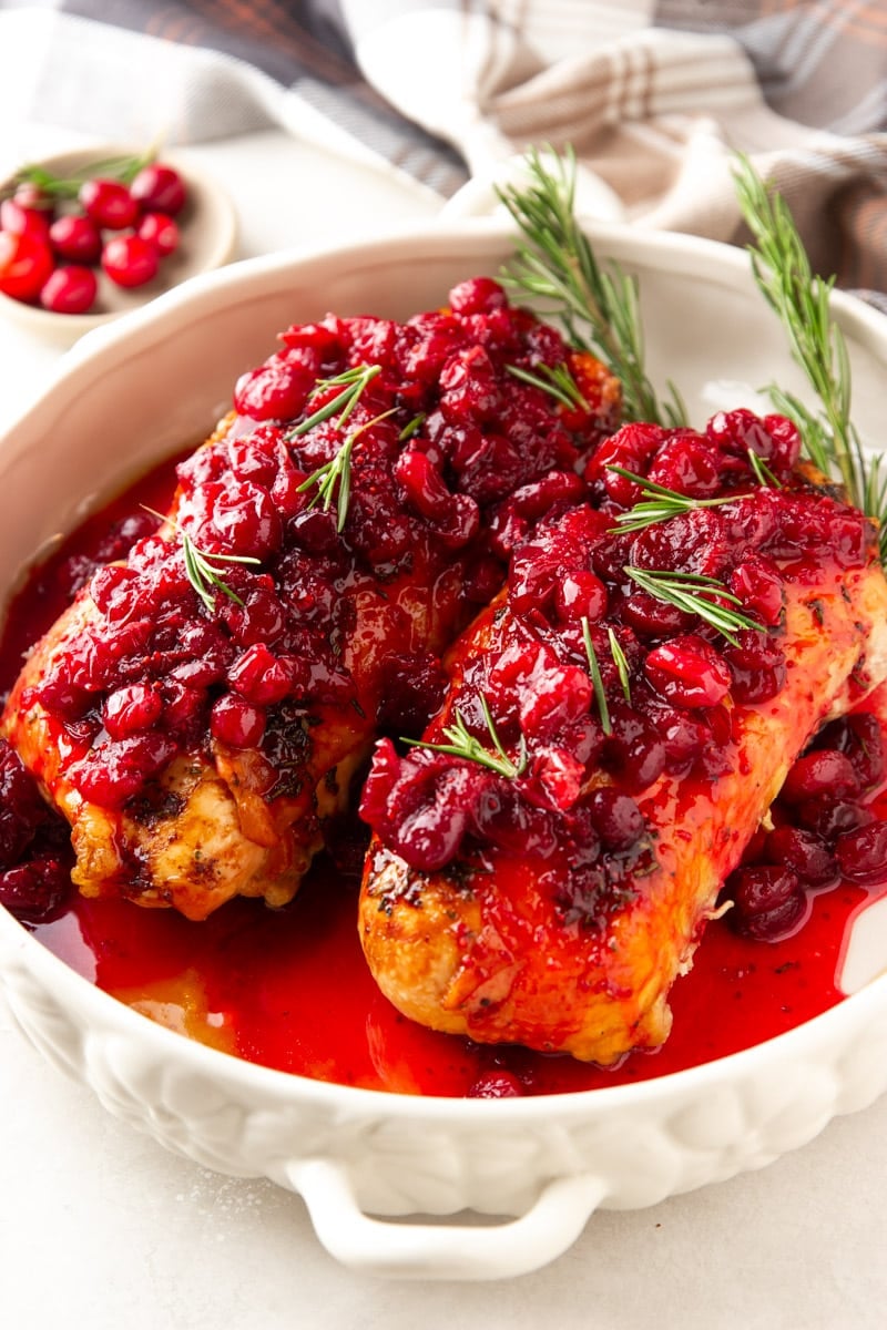 Cranberry glazed turkey breast in a white serving dish, cranberries, and rosemary. 