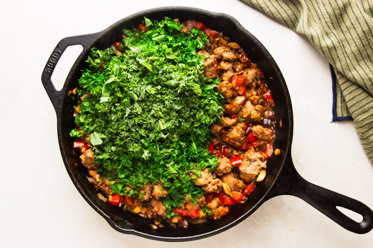 Sausage and veggies with chopped kale in a cast iron pan, and a striped dish towel. 