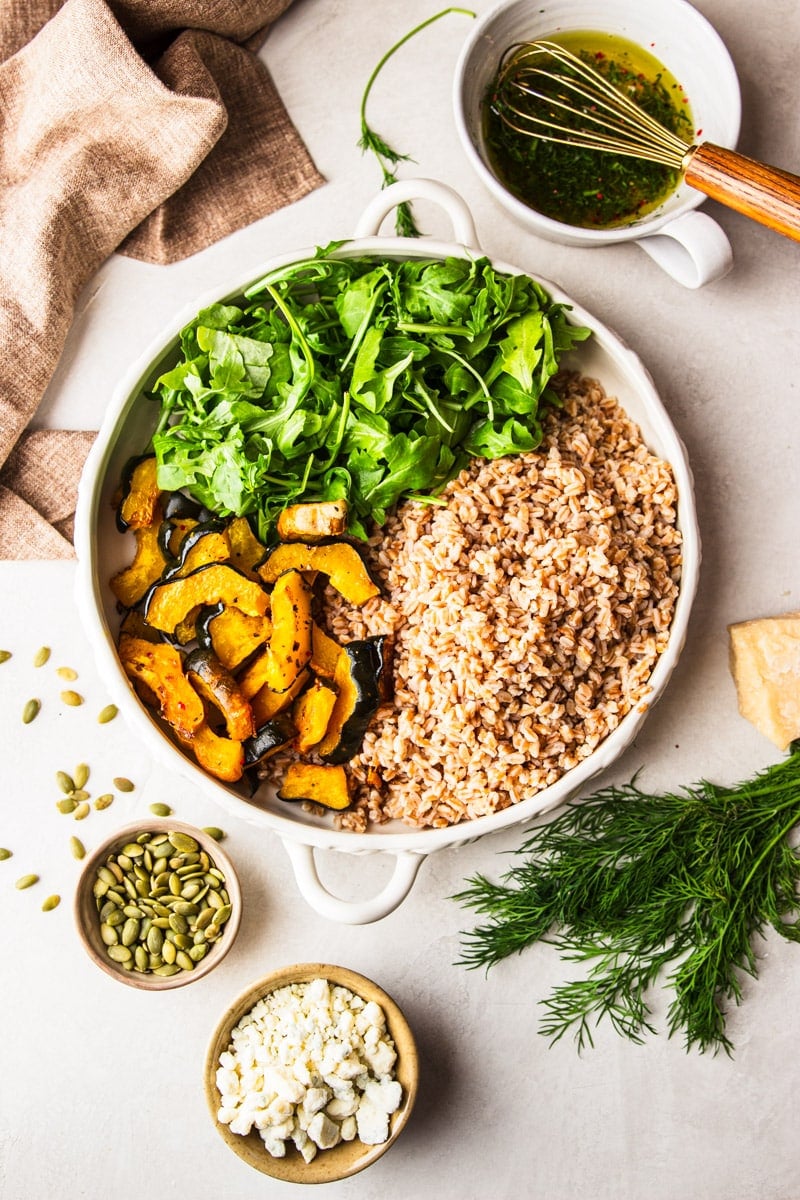 A white bowl of farro, squash and arugula, a bowl of pumpkin seeds, a bowl of dressing with a whisk on top, a block of parmesan cheese, a bowl of crumbled blue cheese, and fresh dill. 