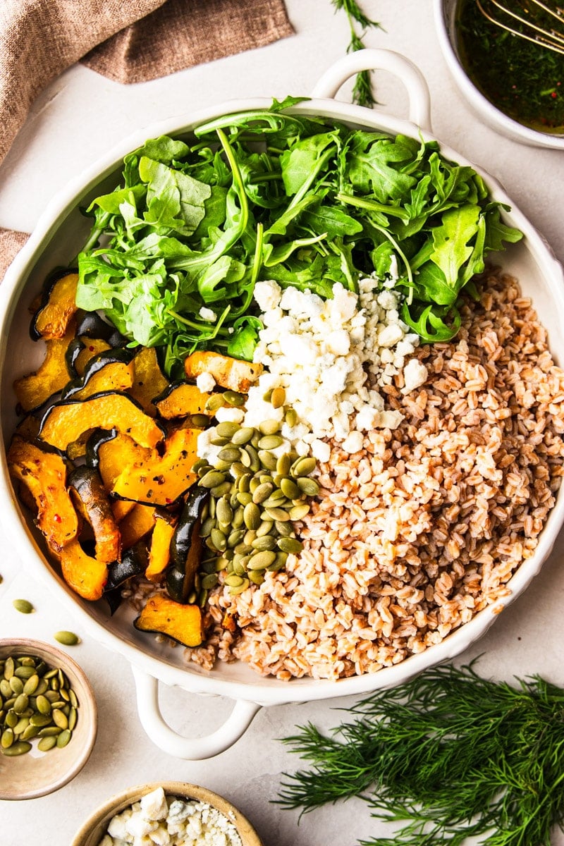 A close up of a bowl of farro, arugula, roasted squash, crumbled blue cheese, pumpkin seeds with a bowl of pumpkin seeds, a bowl of crumbled blue cheese and fresh dill on the side. 