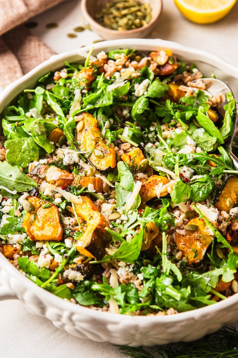 Farro salad with roasted squash in a white bowl with a spoon, bowl of pumpkin seeds, and half a lemon.