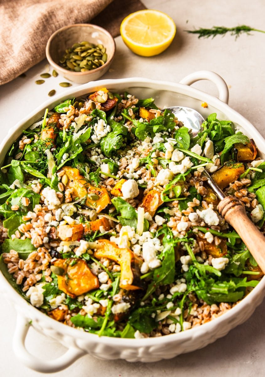 Warm farro salad with roasted squash in a bowl with a spoon, a bowl of pumpkin seeds, half a lemon, and fresh dill. 